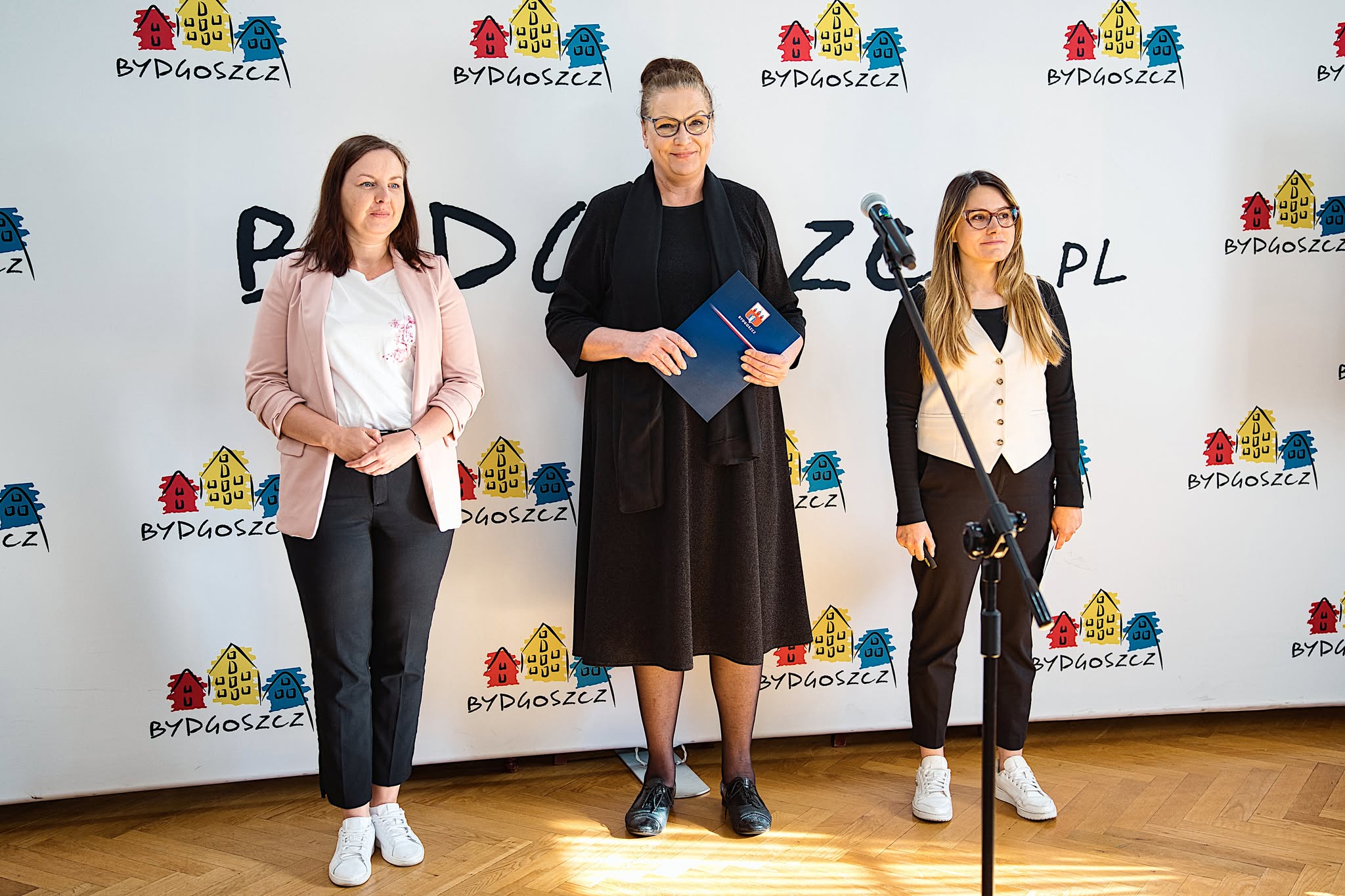 Trzy kobiety stoją obok siebie na tle ścianki z logo miasta Bydgoszczy. Przed nimi ustawiony jest mikrofon.